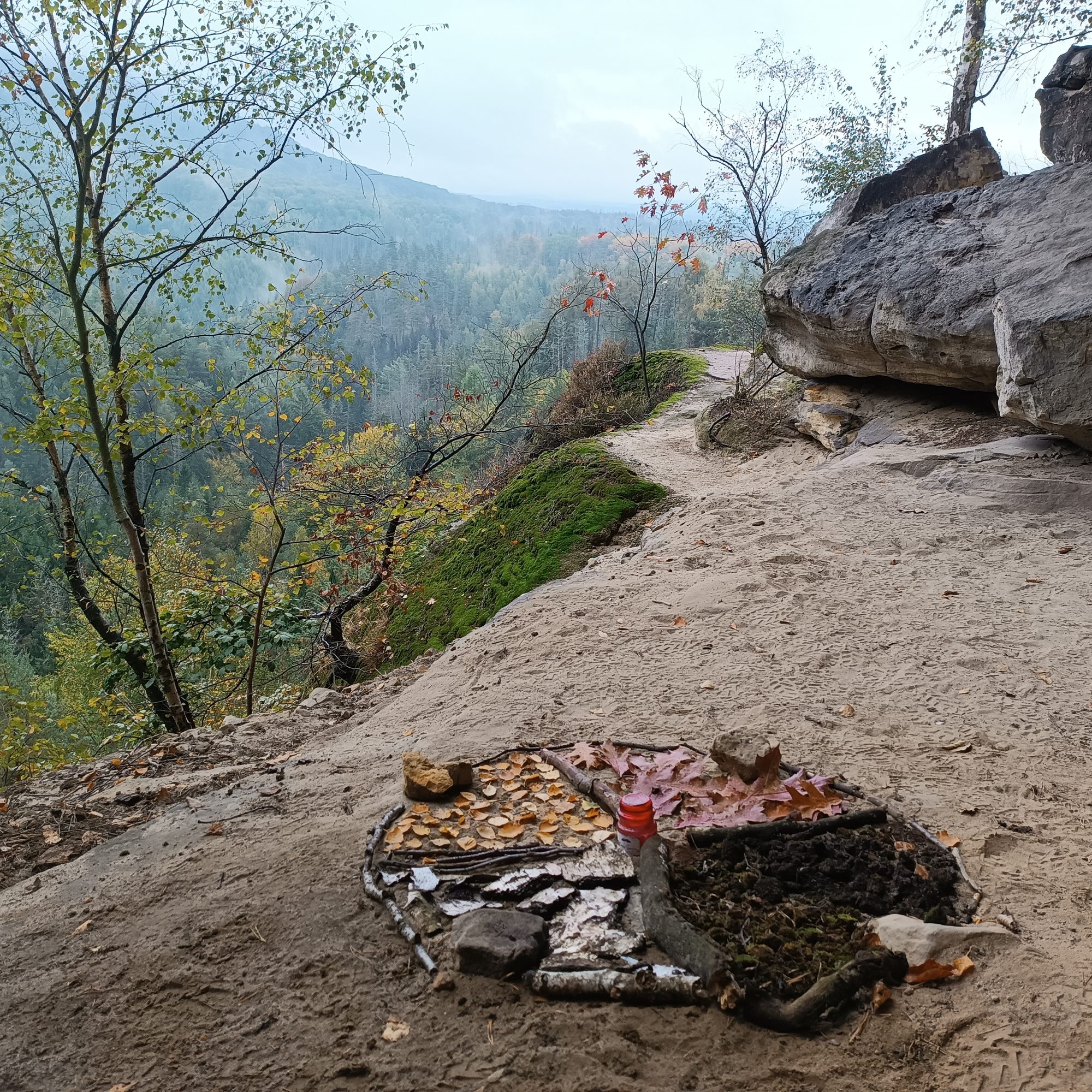 Ein Mandala aus Herbstlaub, Rinde, Erde und Steinen auf einem Felsvorsprung über einem nebligen Wald